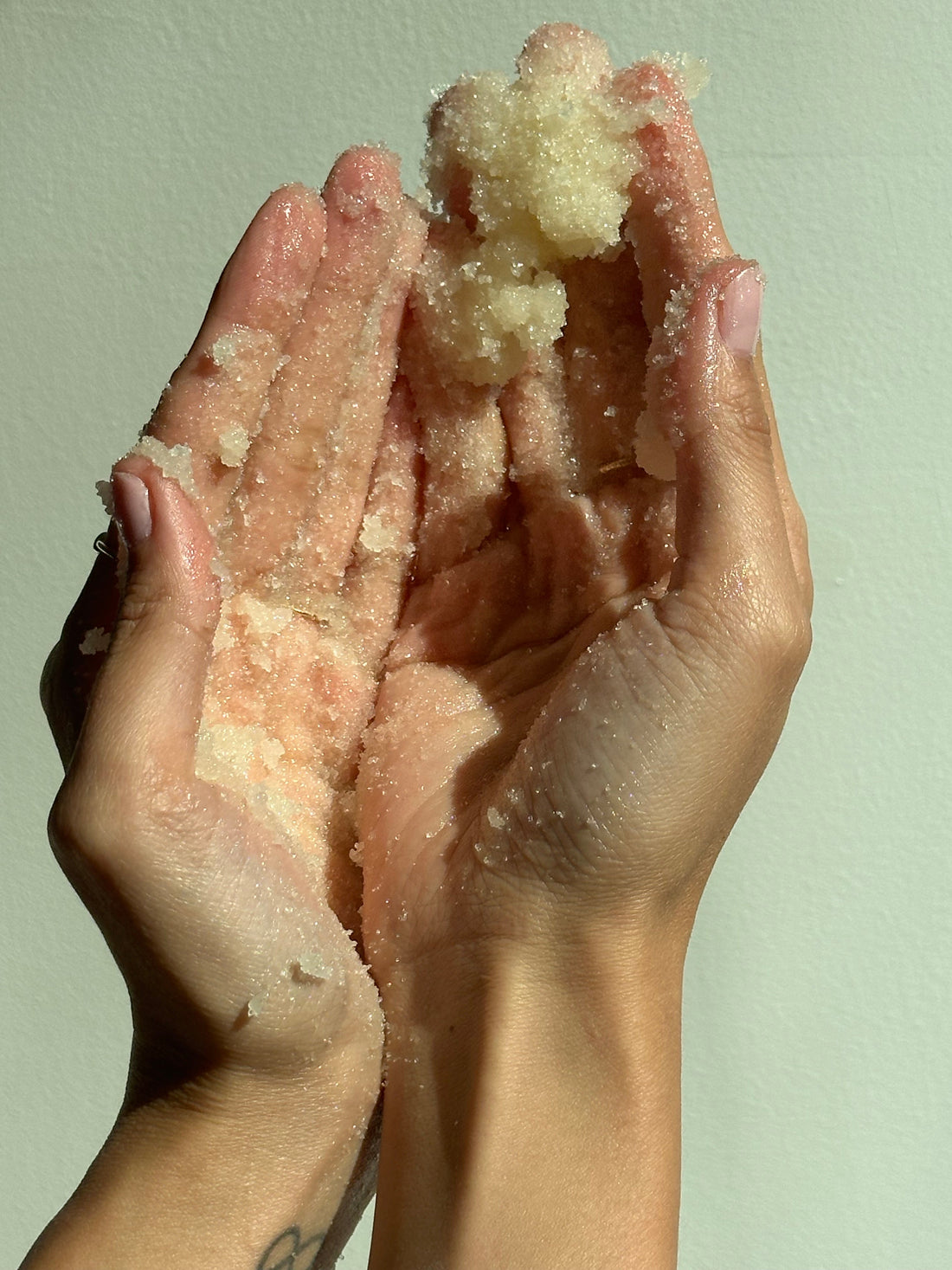 Close-up of two hands holding a scoop of grainy exfoliating scrub against a neutral background.