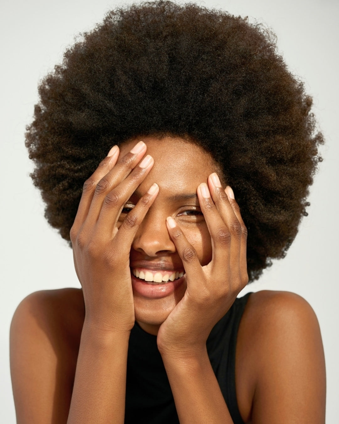 Woman with an afro covering her face with her hands on a plain background