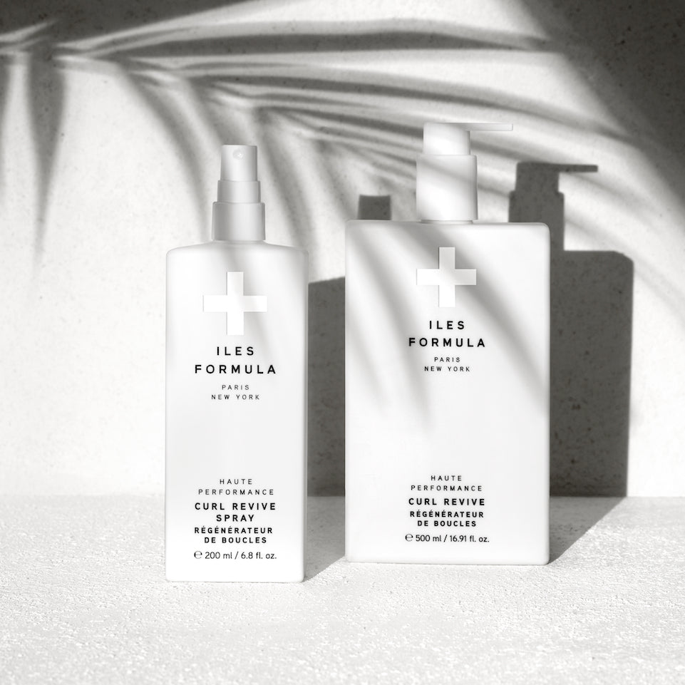 Two white bottles of Iles Formula Haute Performance Curl Revive Spray and Curl Revive, displayed on a textured surface with palm leaf shadows in the background.