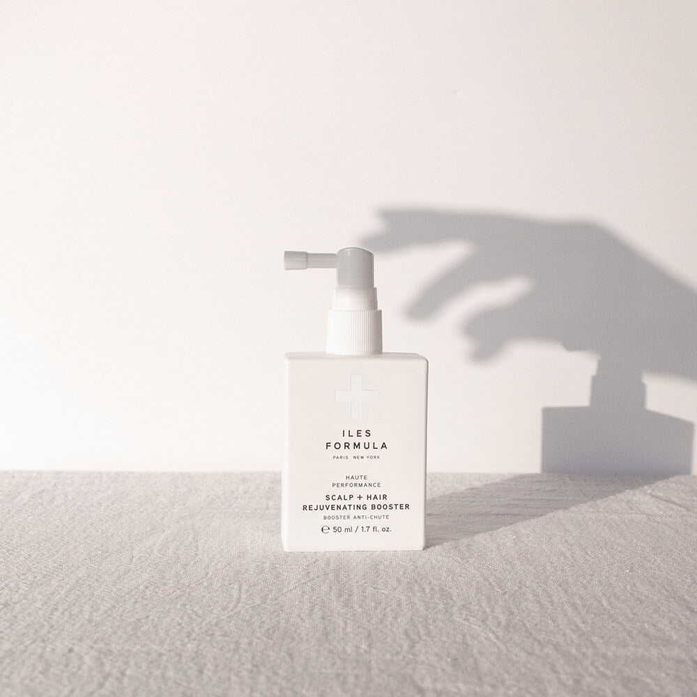 White bottle of Iles Formula Scalp + Hair Rejuvenating Booster with pump dispenser, placed on a textured surface with a hand shadow cast on the wall behind.