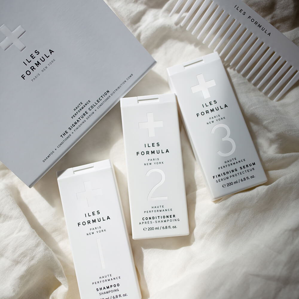 Two white bottles of Iles Formula Haute Performance Curl Revive Spray and Curl Revive, displayed on a textured surface with palm leaf shadows in the background.