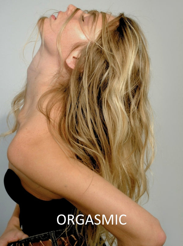 Person with long blonde hair and a black garment on a plain background, with the word 'ORGASMIC' displayed.